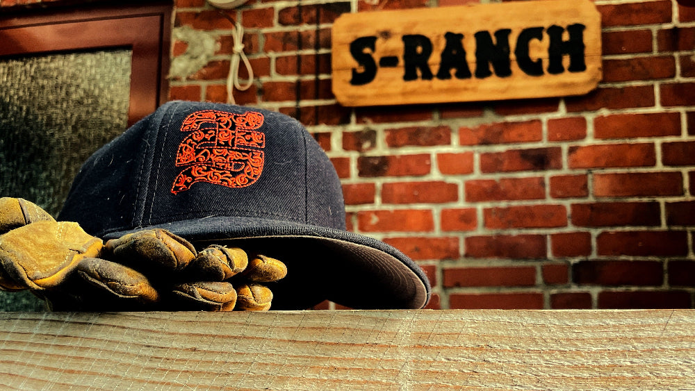A navy blue baseball cap with an orange graphic logo sits on a wooden table, with a pair of tan leather work gloves resting on the brim. A brick wall and a wooden sign reading 'S-RANCH' are visible in the background.